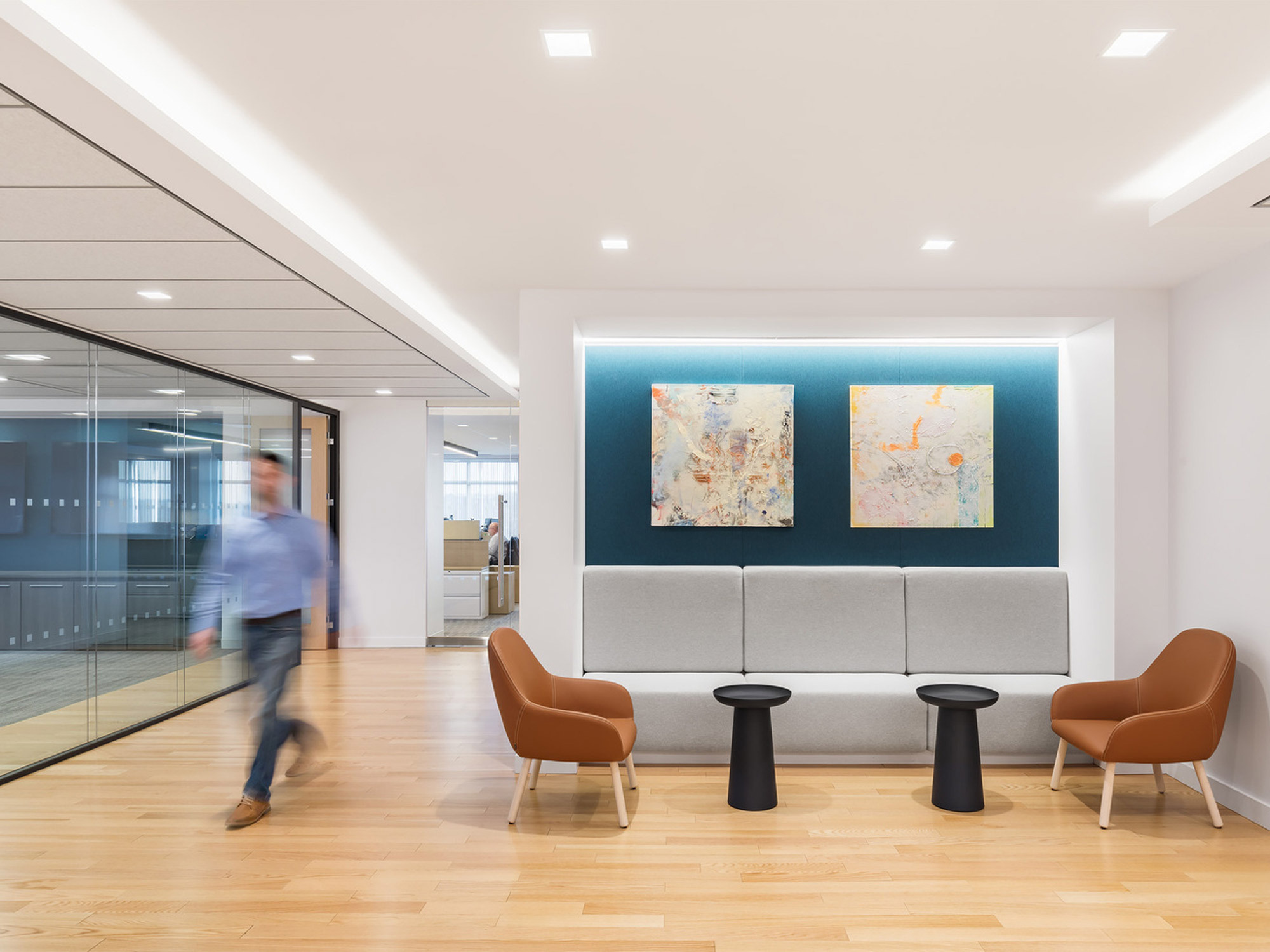 Modern office lobby featuring a neutral color palette with blue accent wall panels, two abstract paintings, sleek grey seating, and warm wooden flooring. Glass partitions offer a glimpse into the adjacent space, highlighting the open-concept design, while recessed lighting provides ambient illumination.