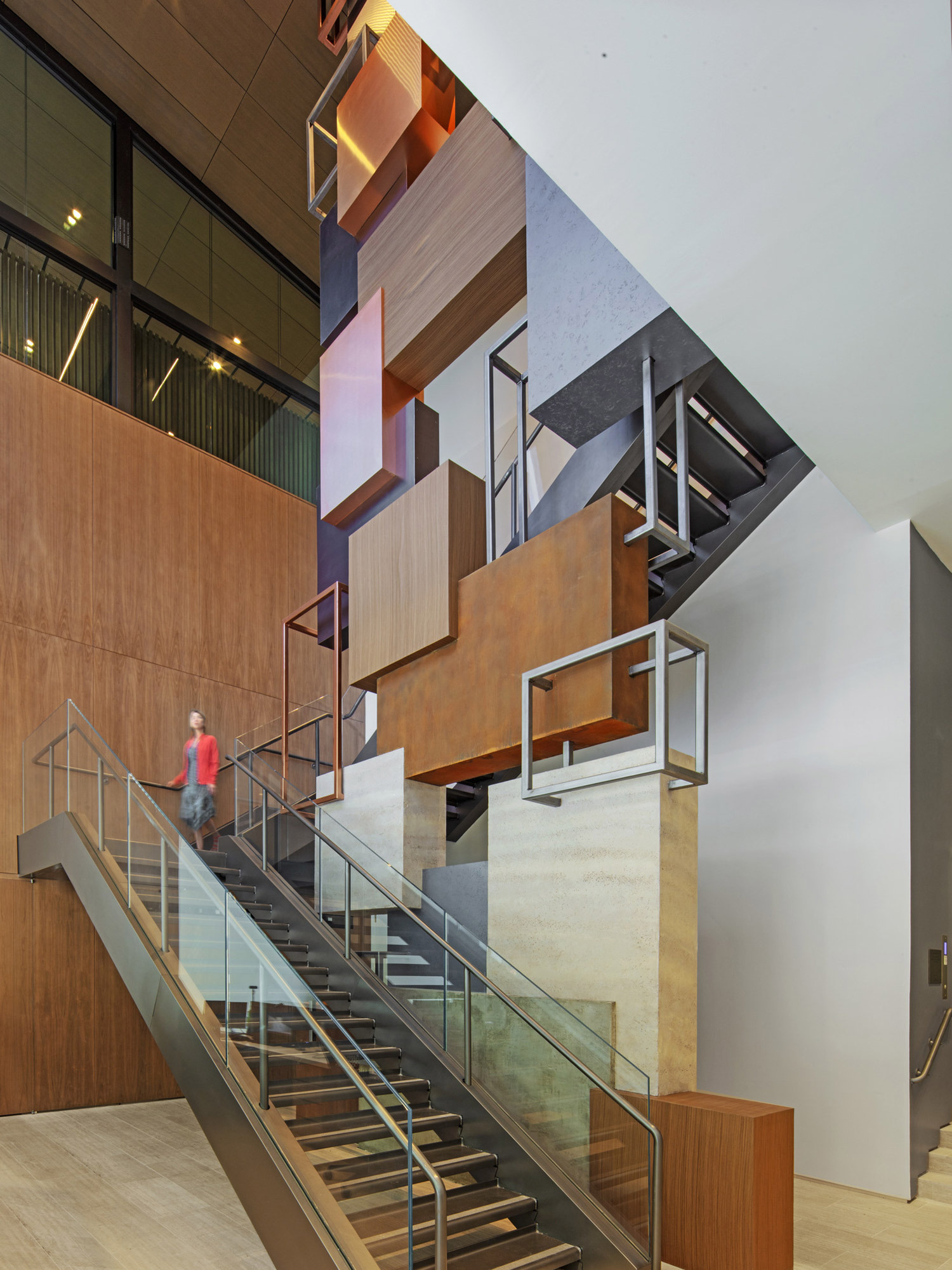 Modern staircase intersects warm wooden panels and a large abstract painting, accented by geometric pendant lights and glass balustrades, providing an engaging vertical flow within a contemporary space.