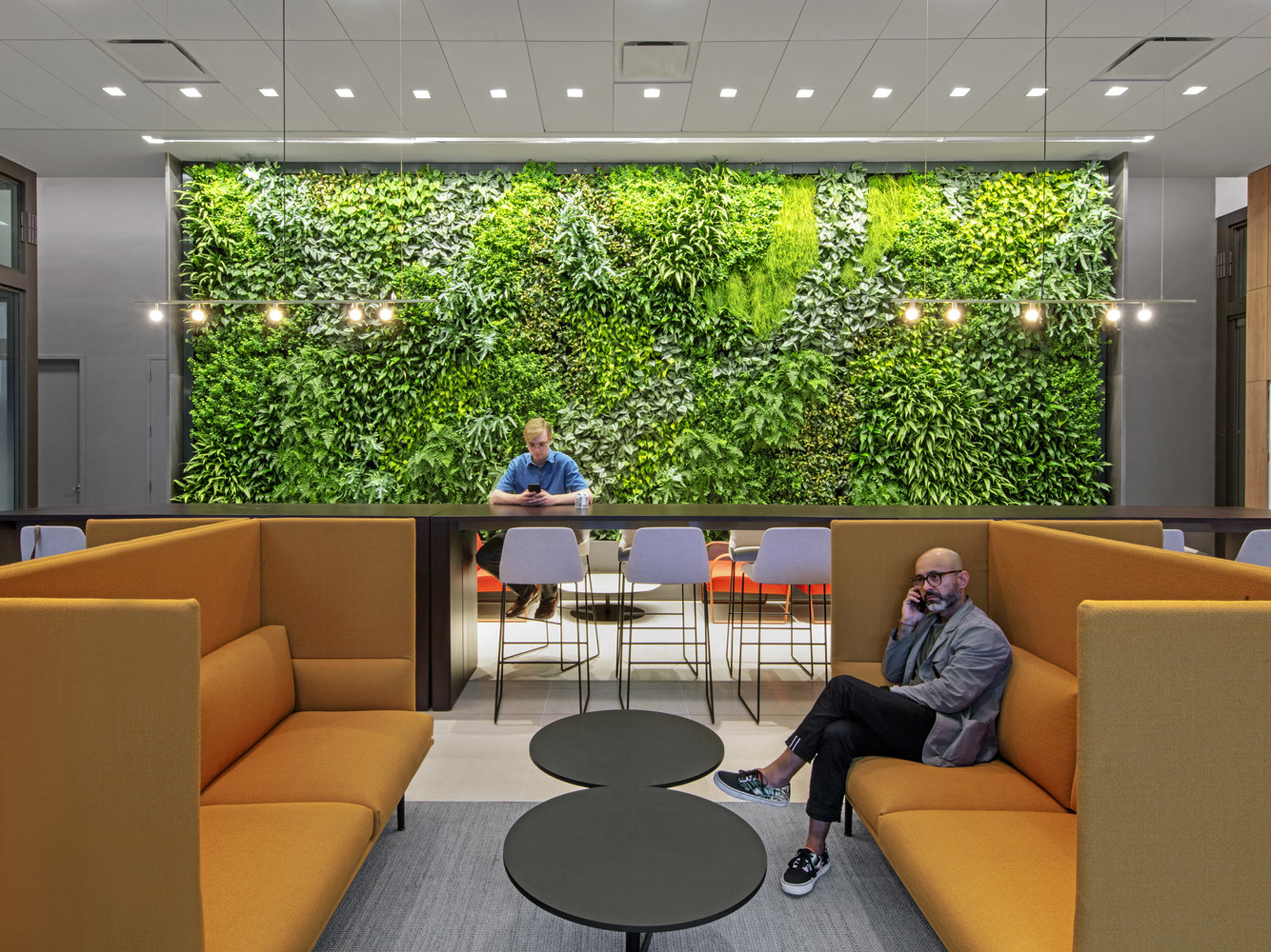 Modern office break area with vibrant, upholstered orange modular seating and a central black coffee table. Two people converse casually, flanked by a lush living green wall, providing a biophilic design element to the contemporary, well-lit space.