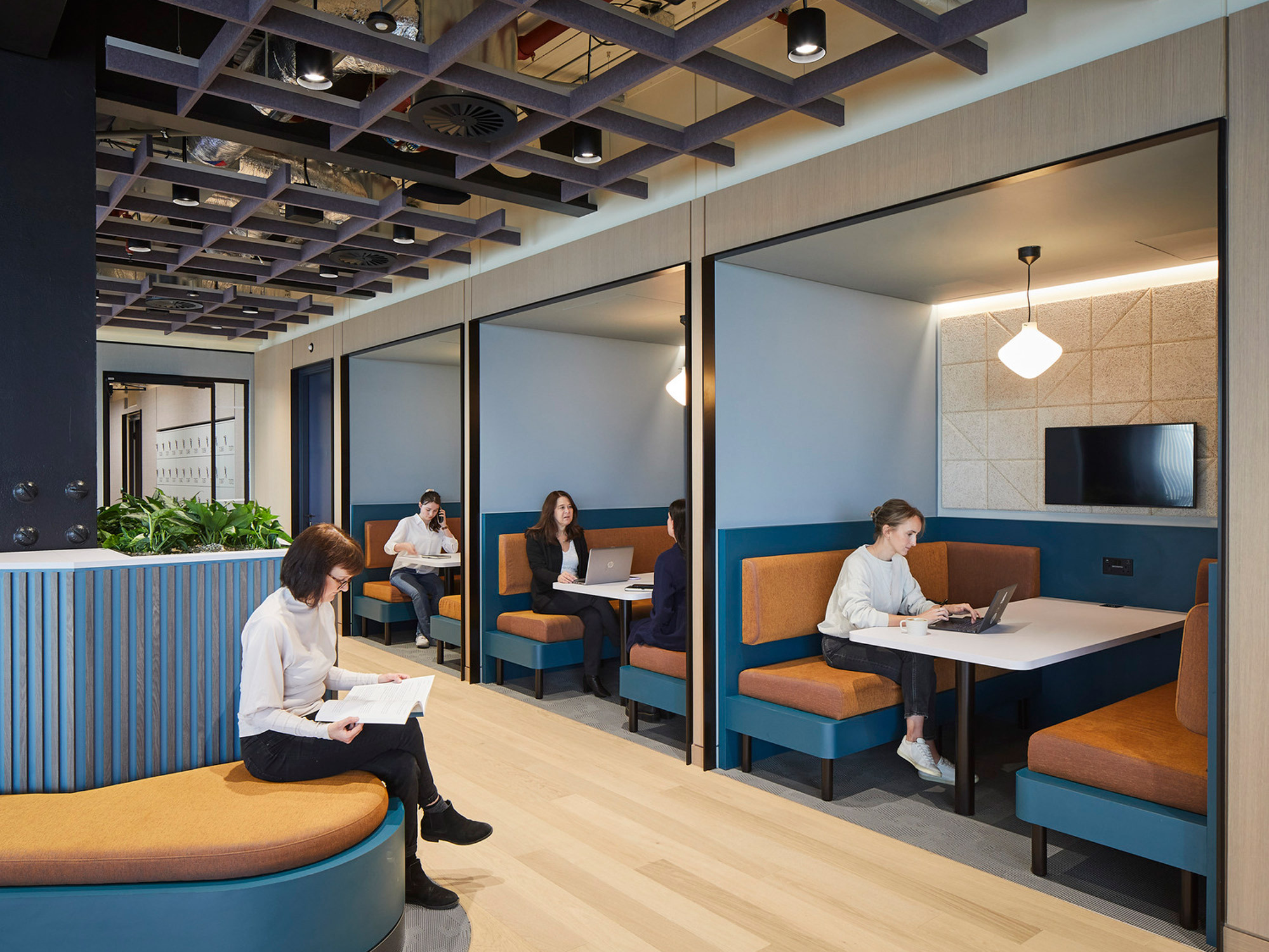 Modern office space featuring private blue and terracotta booths with integrated seating and tables, set against paneled walls. Overhead, an exposed ceiling adds an industrial touch, complemented by warm pendant lighting, providing functionality and aesthetic appeal for collaborative work environments.