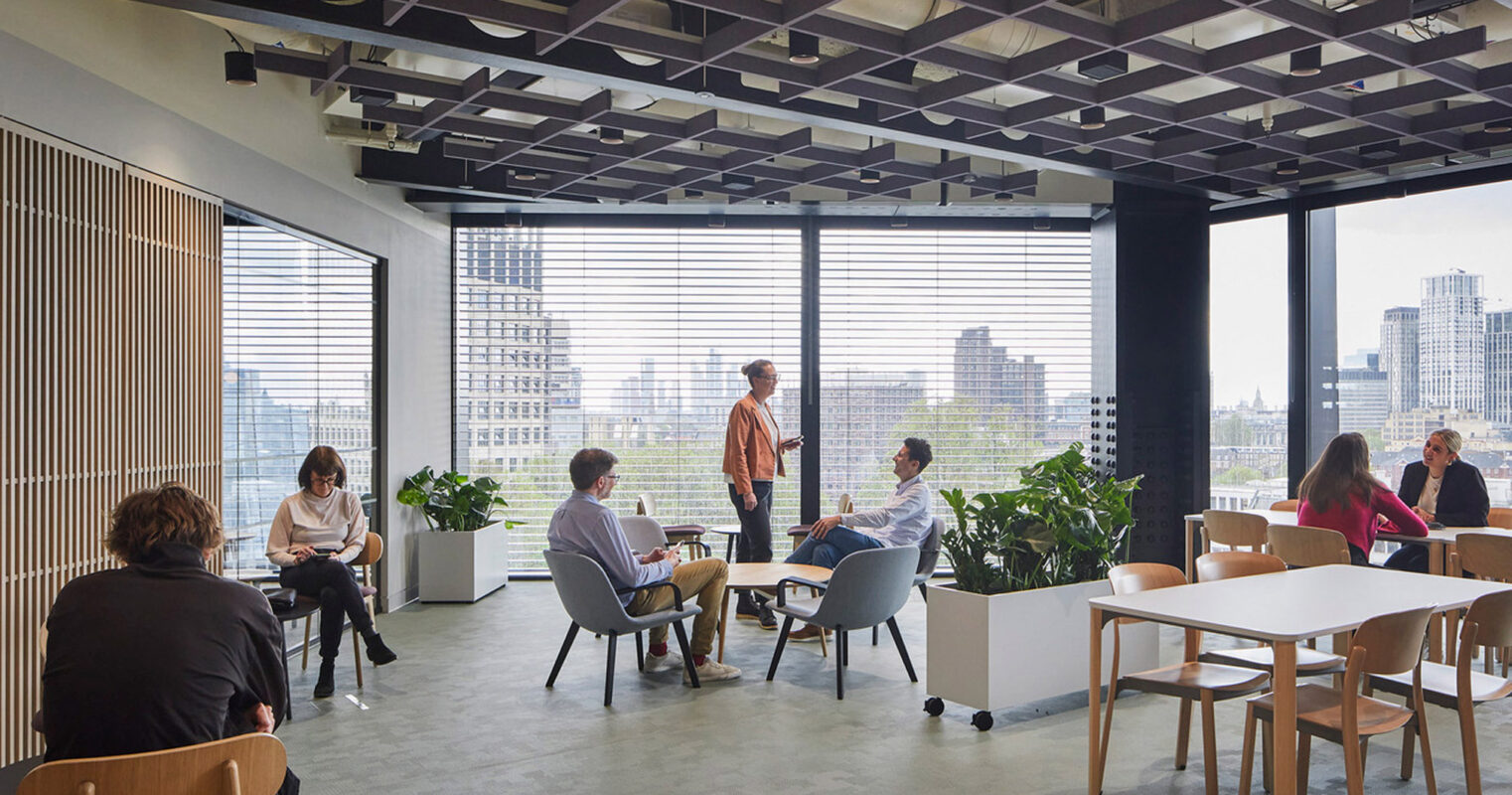 Open-plan office space with natural light, featuring a mix of standing and seated workstations, minimalist furniture, and indoor greenery, overlooking an urban skyline. Vertical slats partition the area while preserving a sense of openness.