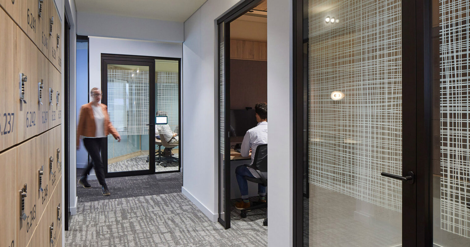 Contemporary office space featuring glass partition walls with embedded wire mesh, modern blue cabinetry with numbered lockers, and gray textured carpeting. Design promotes transparency and efficient use of space.