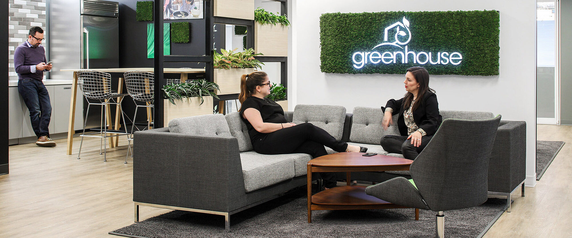 Modern office lounge with plush gray sofas and wooden coffee table. Green wall with company logo enhances biophilic design, while wood accents and clean lines convey contemporary style. Natural light and plants create a welcoming atmosphere for collaboration.