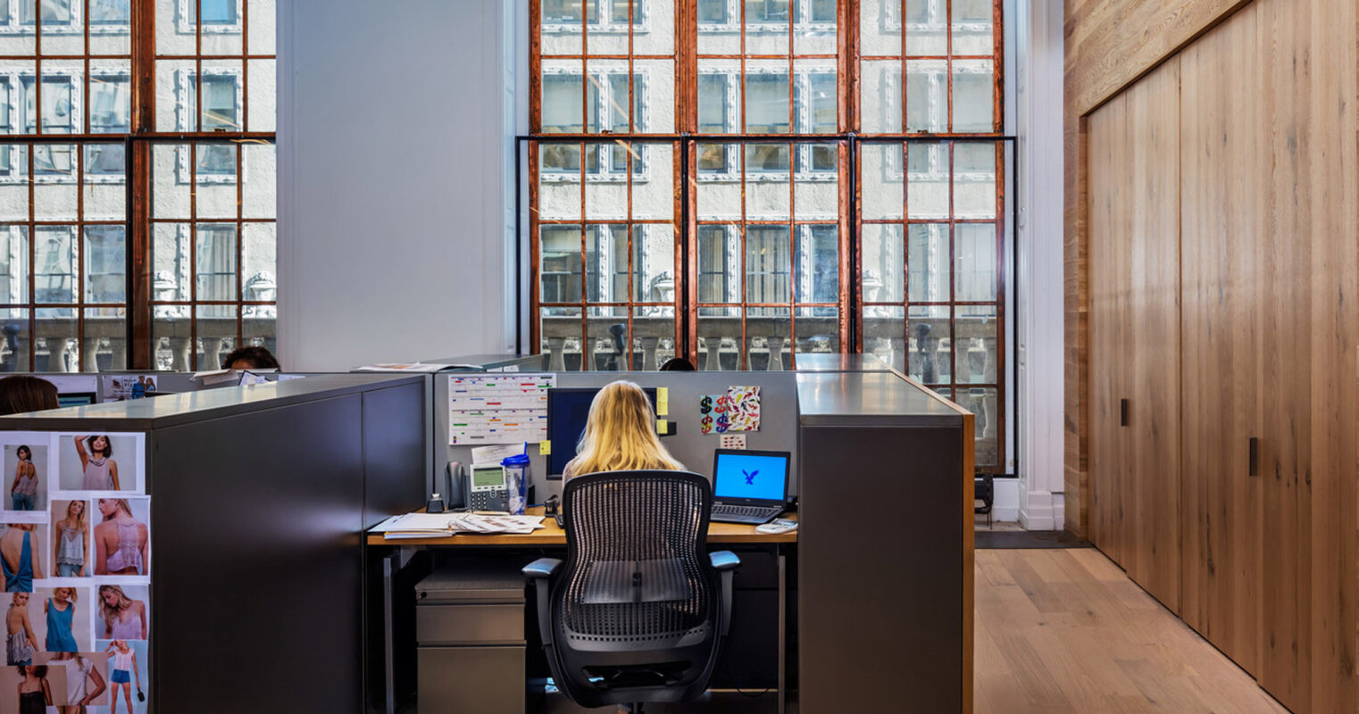 Open-plan office space featuring a workstation with a seated employee focusing on a laptop. Exposed brick façade with large windows allows natural light, complementing the warm tones of the wooden wall cladding and flooring. A personal mood board adds a touch of individuality to the professional setting.