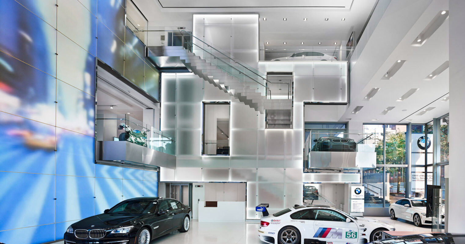 Spacious, modern car showroom with high ceilings, featuring a prominent, geometric glass and metal staircase leading to an upper level. Gleaming luxury vehicles are arranged below on a polished concrete floor, flanked by sleek, white support columns. Dynamic blue ambient lighting enhances the futuristic ambiance.