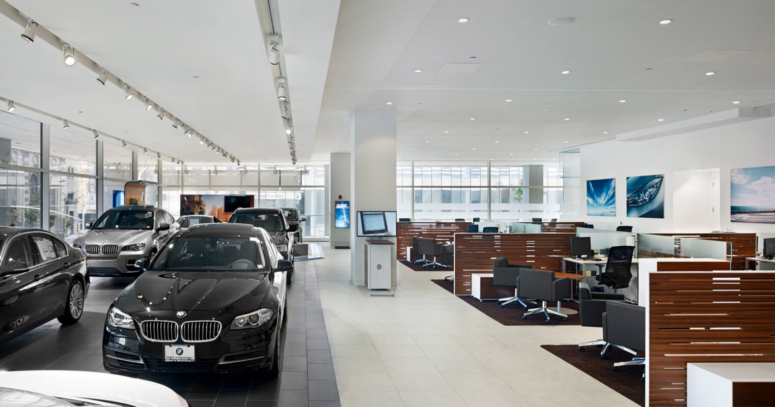 Spacious car showroom featuring sleek, modern design with white flooring and ceiling, ample natural light, and rows of high-end vehicles alongside elegant wood-paneled cubicle workstations.