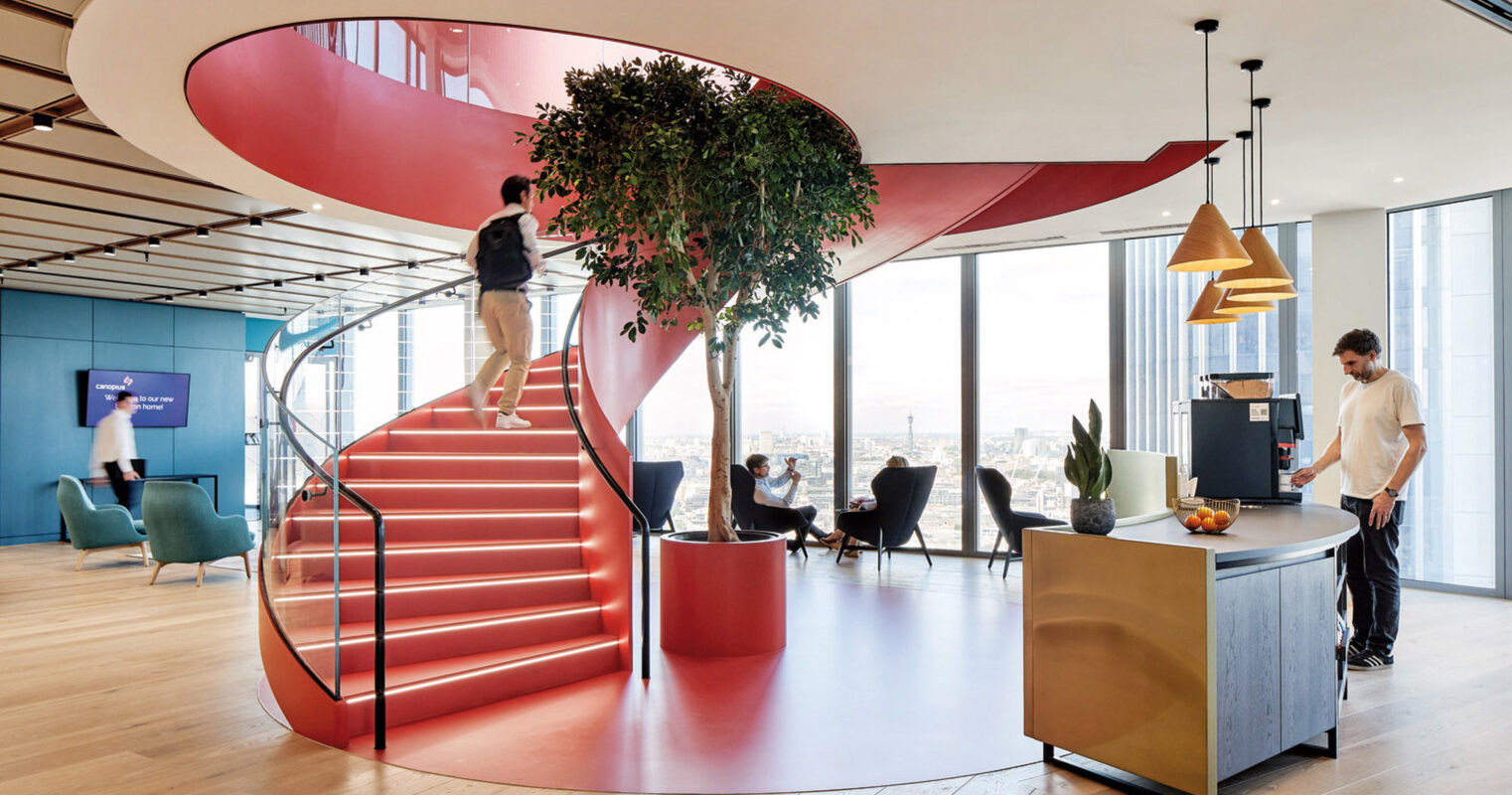 Modern office space with an open-plan layout featuring bold red spiral staircase, central green tree, vibrant blue wall panels, and eclectic furniture that encourages interaction and collaboration. Natural light floods the area, creating a dynamic, energizing workplace.