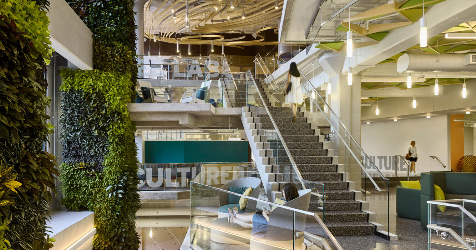 Modern office interior featuring a grand staircase with glass balustrades next to a verdant living wall. The space is illuminated by sculptural ceiling lights reflecting on reflective surfaces, emphasizing the blend of natural elements with contemporary design.