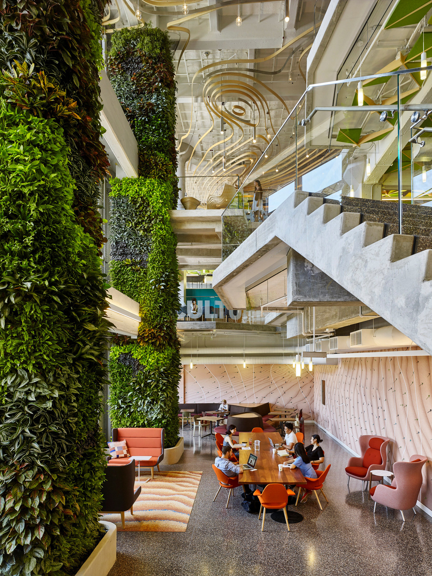Vertical garden flanks a glass-walled interior space with a floating concrete staircase. Exposed ceiling ducts contrast with plush seating and warm wood tones, promoting a blend of industrial and organic design elements.