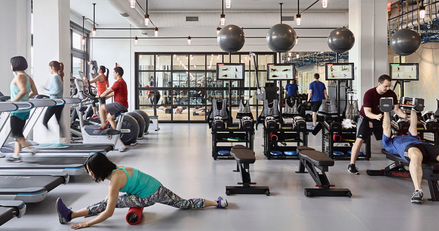 Modern gym interior featuring a variety of exercise equipment, mirrored walls reflecting treadmill users, and an open floor plan with ample natural light. The space is characterized by a clean, industrial aesthetic with exposed ductwork and a contrasting mix of dark and light finishes.