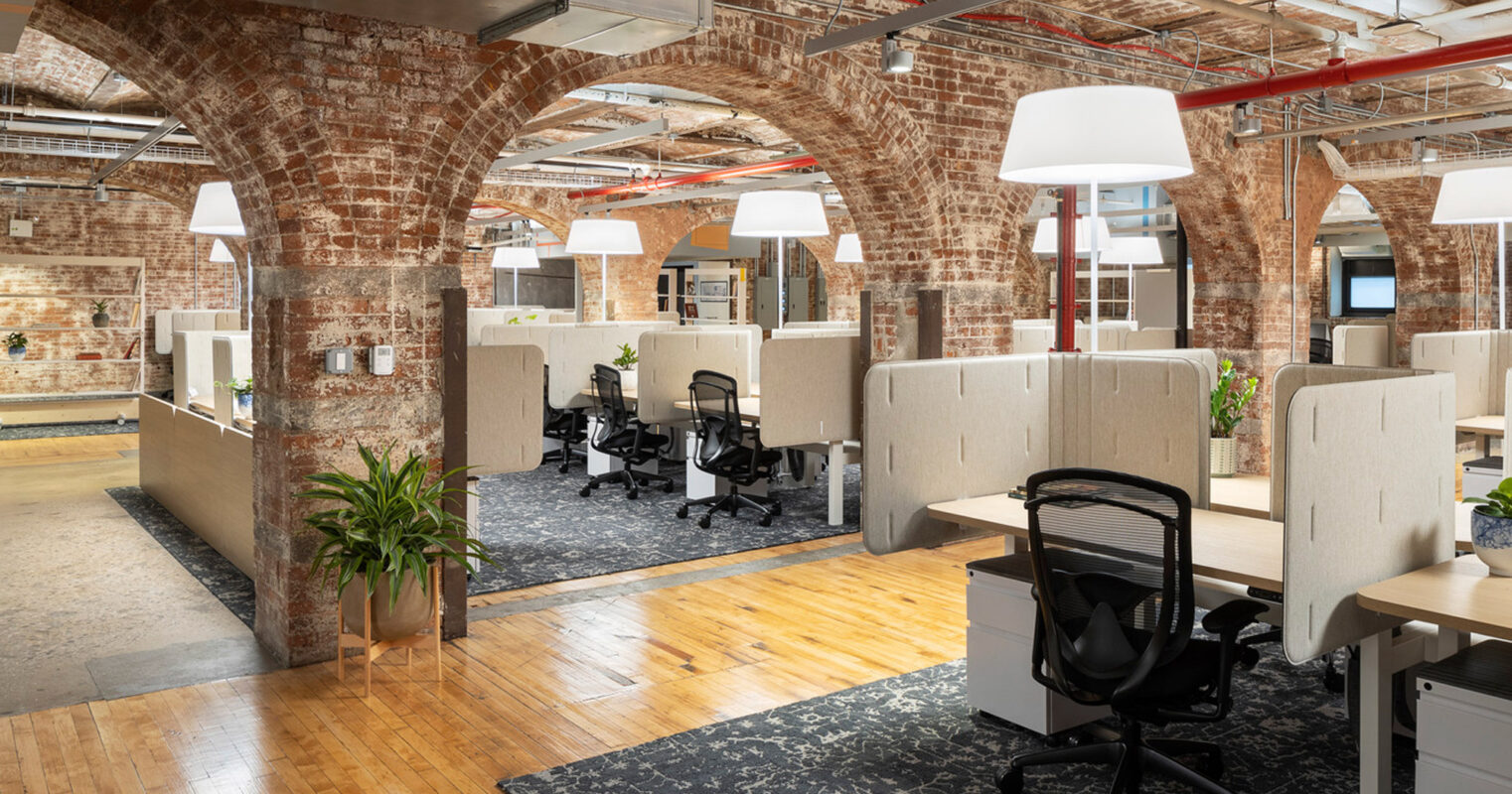 Modern workplace in a historic loft-style building with exposed brick walls and arched ceilings. Features include hardwood flooring, contemporary cubicles, pendant lighting, and strategically placed greenery, creating a fusion of industrial charm and ergonomic design.