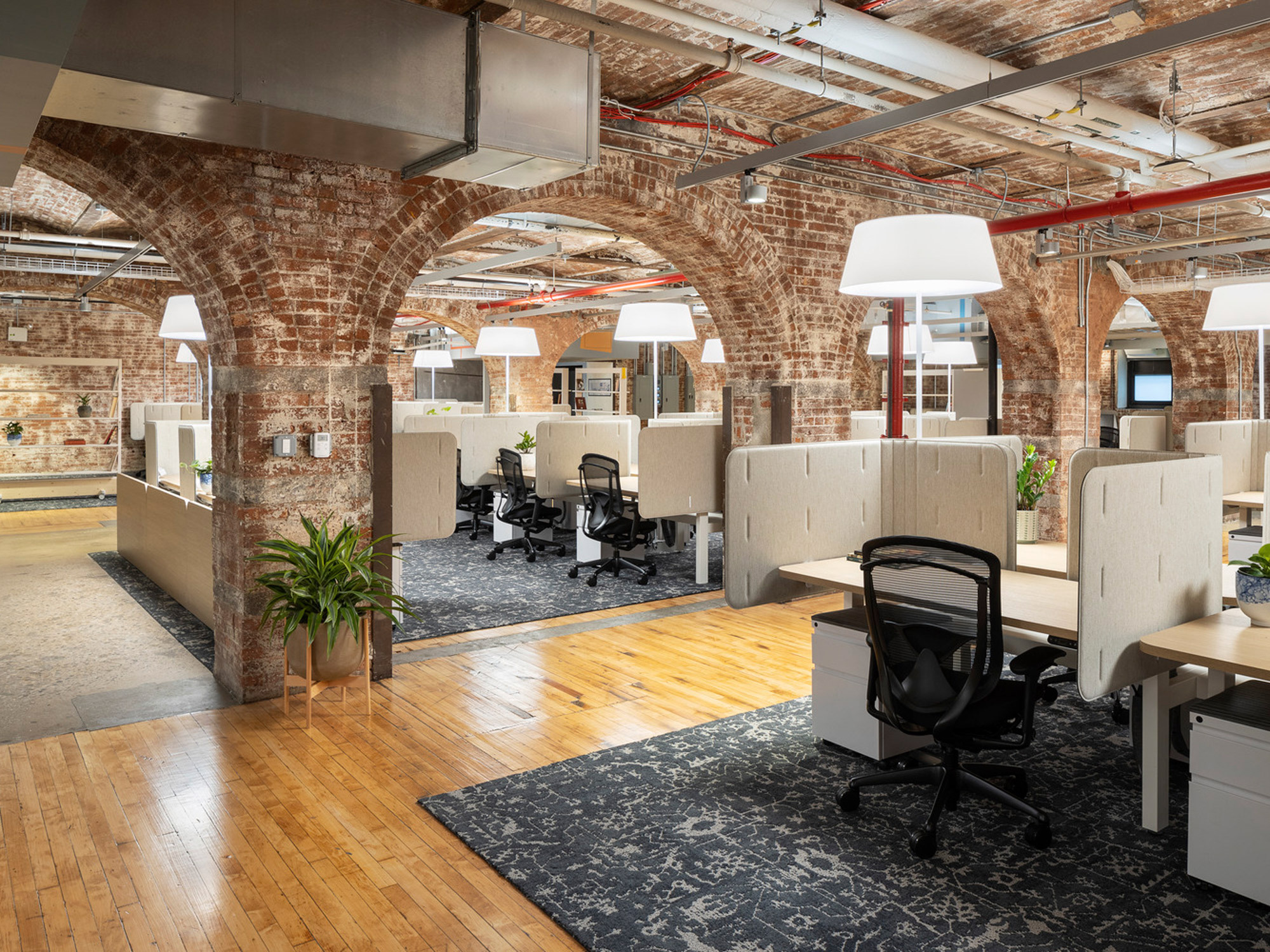 Modern workplace in a historic loft-style building with exposed brick walls and arched ceilings. Features include hardwood flooring, contemporary cubicles, pendant lighting, and strategically placed greenery, creating a fusion of industrial charm and ergonomic design.