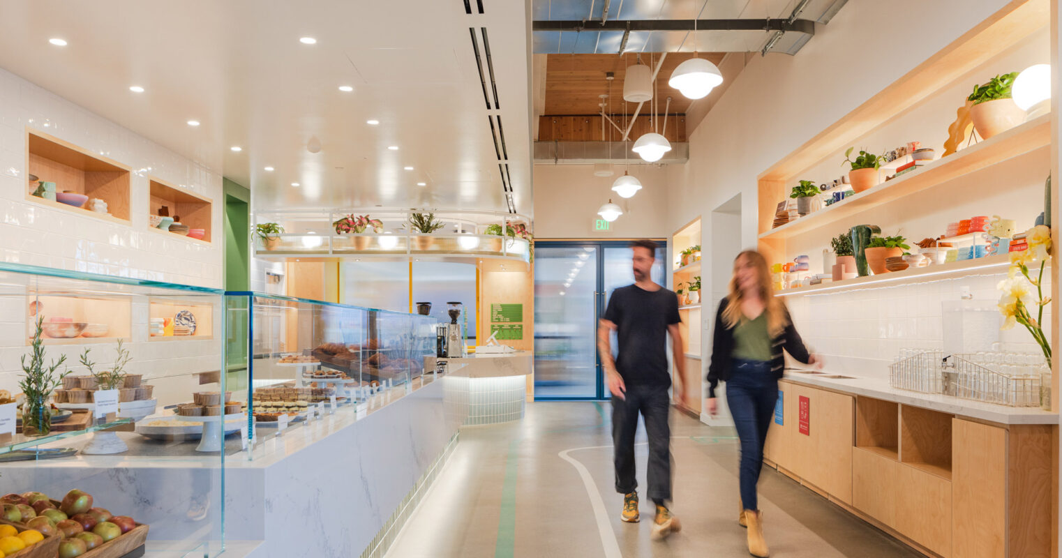 Open-concept café interior featuring natural wood finishes, recessed lighting, and custom shelving displaying fresh produce. A central glass partition accents the modern, inviting space.