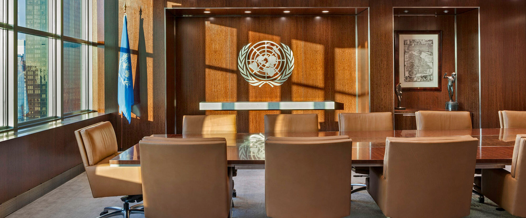 Modern conference room with floor-to-ceiling windows providing ample natural light, complementing the rich wooden wall panels and sleek, rectangular table. Brown leather swivel chairs encircle the table, while the United Nations emblem commands attention on the focal wall.