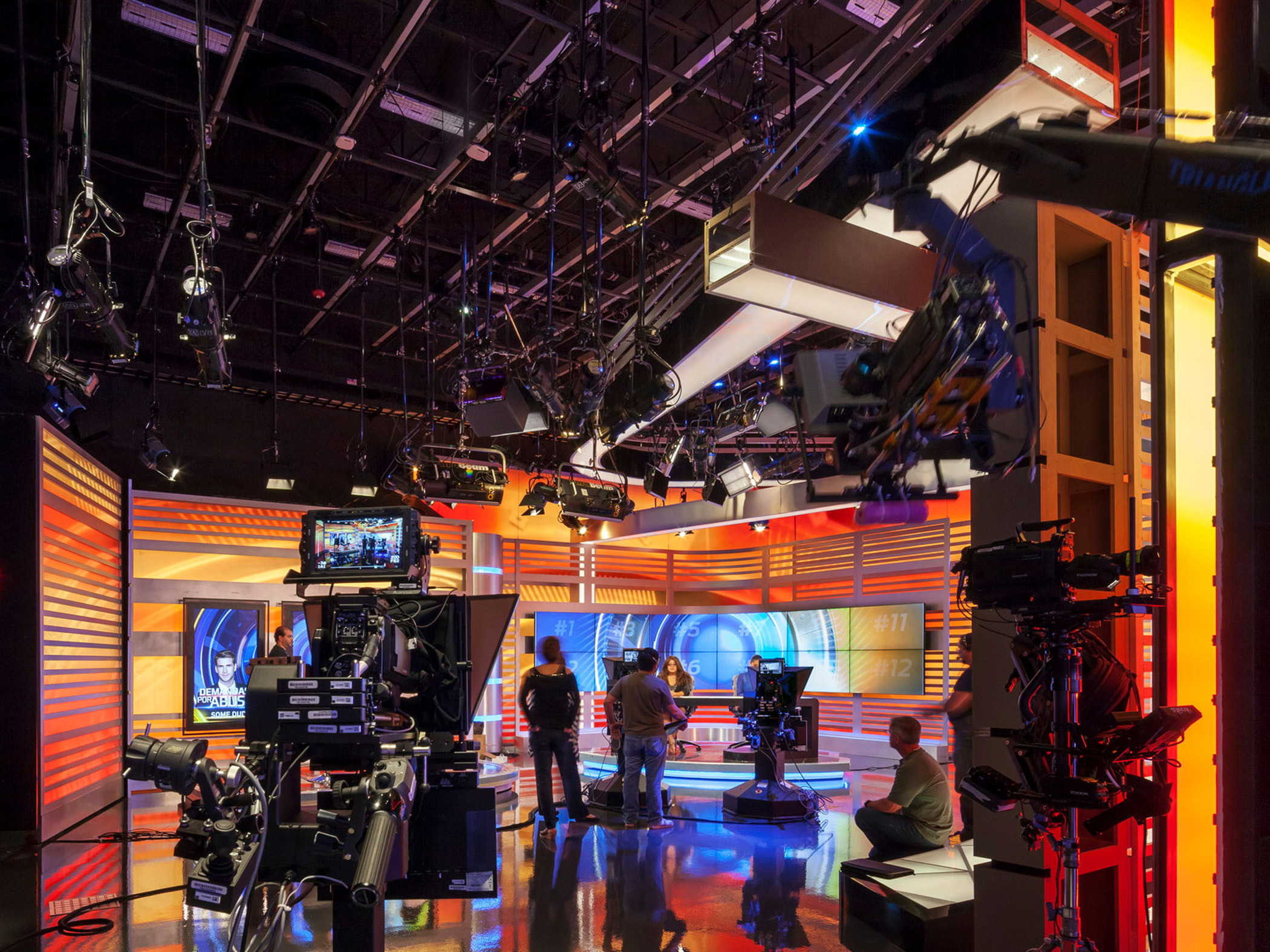 Vibrant television studio interior featuring a dynamic, multilayered set with integrated orange LED lighting. Sleek contemporary lines and geometric patterns define the space, accentuated by high-tech broadcast equipment and cameras poised for live production.
