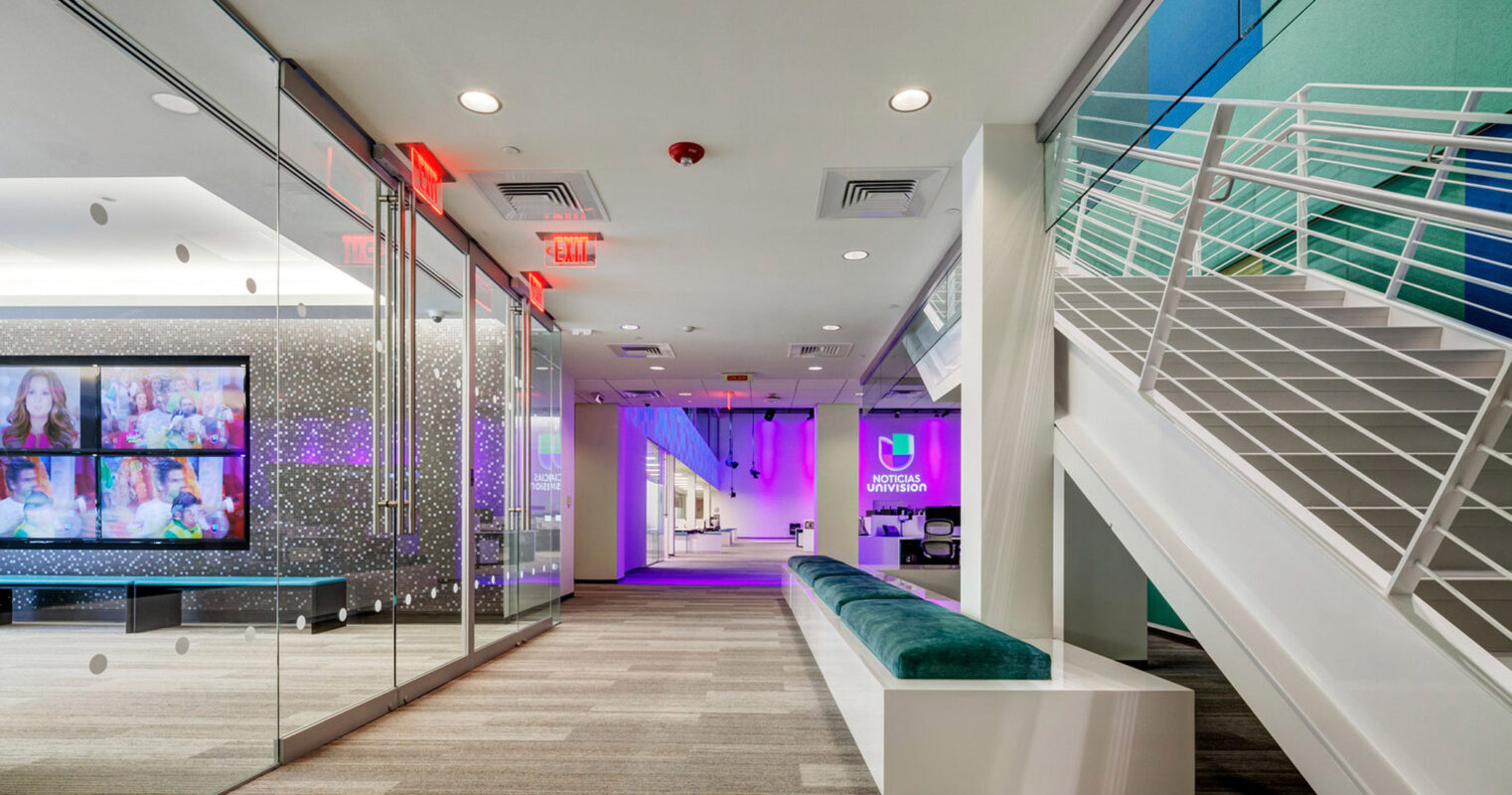 Modern office lobby with vibrant purple accent wall, sleek white staircase with transparent balustrades, and elongated teal bench seating. A multimedia display panel anchors the space, with dynamic lighting and a glass partition reflecting the contemporary design aesthetic.