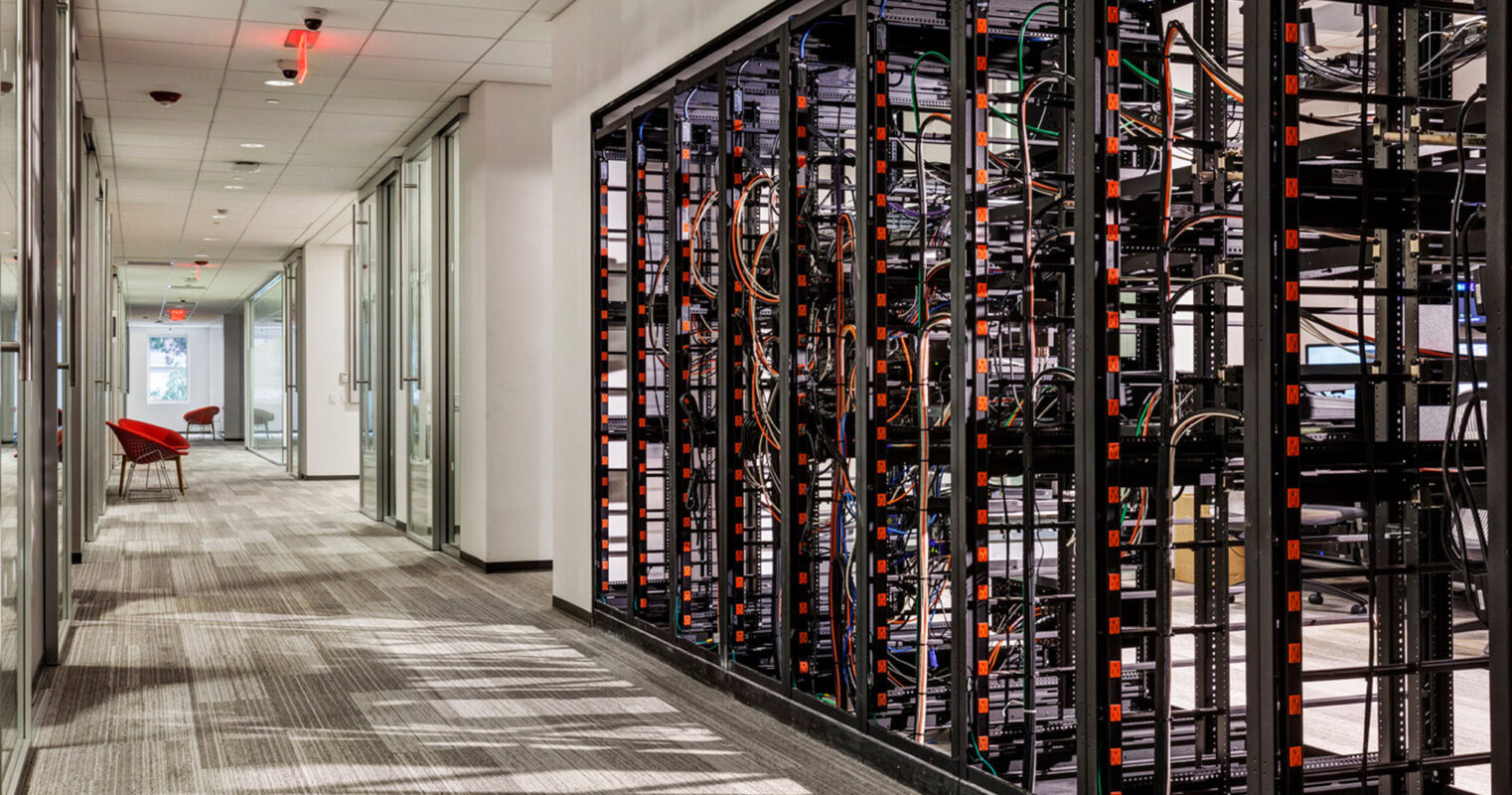 Sleek corporate hallway with a striking contrast between technology and design, featuring an open framework server rack on the right, patterned gray carpeting, and a pop of color from a red chair.