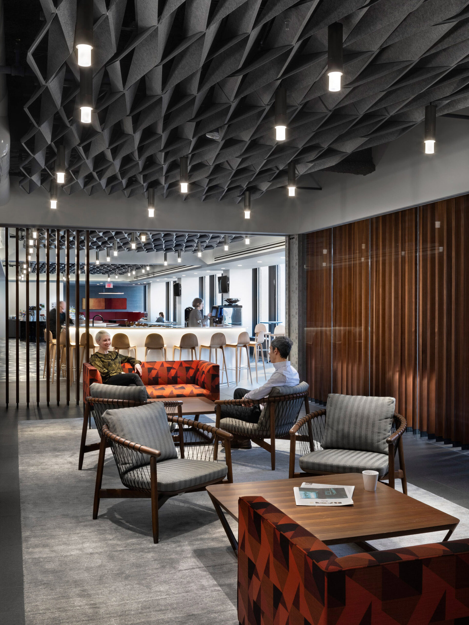 Modern office lounge with geometric acoustic ceiling panels, pendant lighting, and a mix of wooden elements and textured upholstered seating, fostering a contemporary and inviting ambiance.