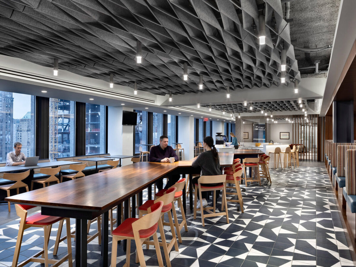 Modern office cafeteria with geometric black and white tiled floor, exposed undulating ceiling structure, and pendant lighting. Communal wooden tables with red chairs accent the space, framed by large windows overlooking an urban skyline.