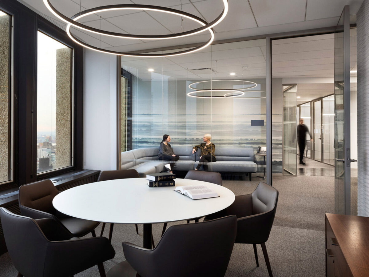 Modern office lounge area featuring sleek, circular overhead lighting, a grey-toned upholstered seating arrangement, and large windows providing ample natural light against an urban backdrop. The space promotes collaboration with a central white round table and dark chairs.