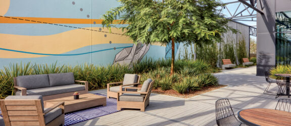 Modern rooftop terrace with industrial touches featuring wooden deck flooring, lush green potted plants, and comfortable seating arranged for social interaction. A geometric mural adds a pop of color to the urban oasis.