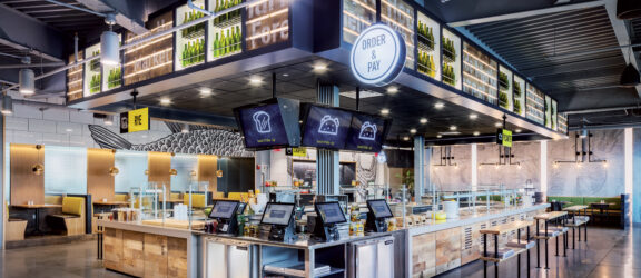 Modern fast-casual restaurant interior featuring an expansive order counter with digital menus, sleek black pendant lighting, and white subway tile backsplash. Organized seating zones and abundant use of natural wood tones complement the industrial chic vibe exemplified by exposed ductwork.