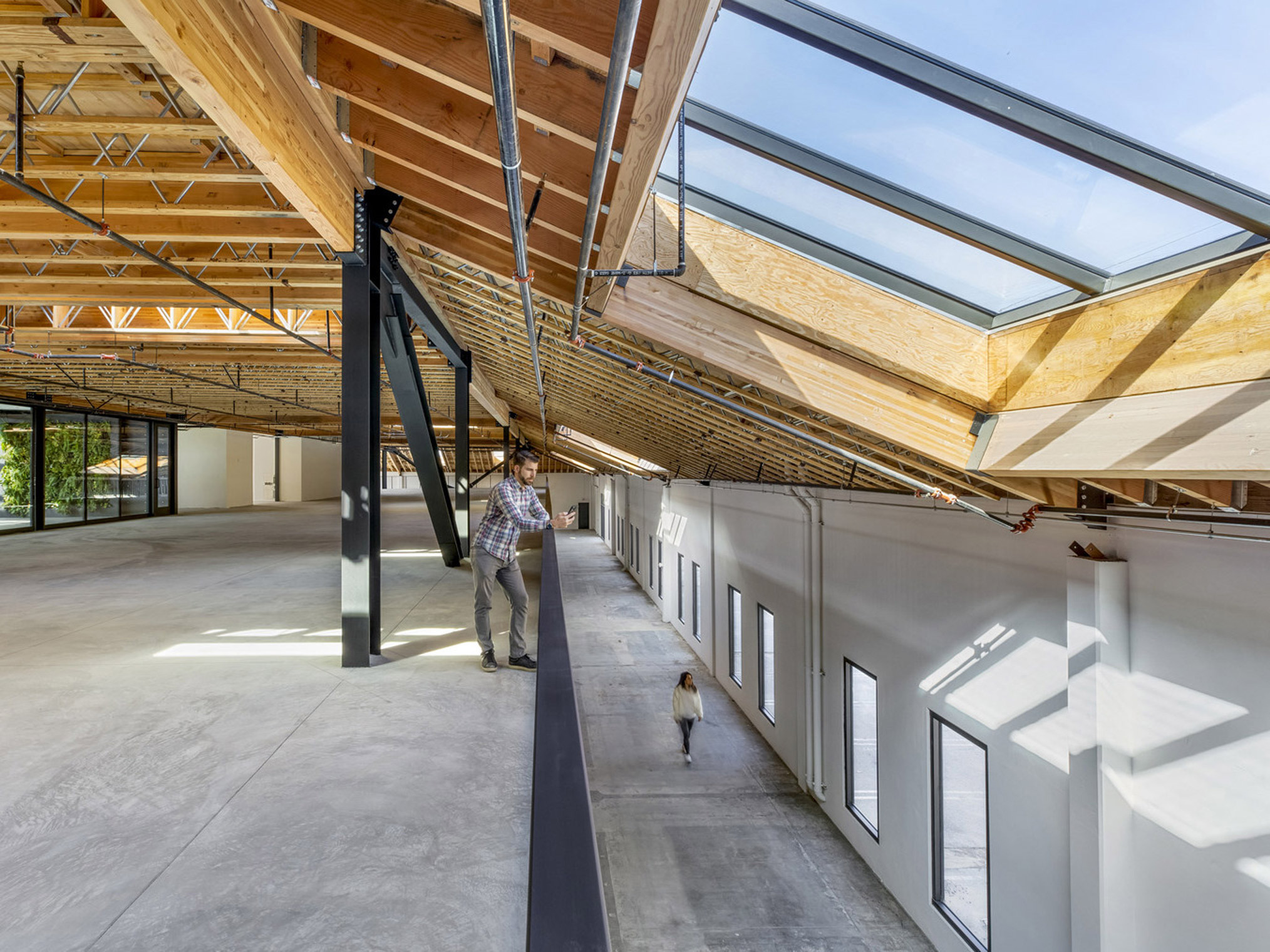 Open-plan office space with exposed wooden beams and diagonal steel braces. Natural light floods in through clerestory windows, highlighting polished concrete floors where a person strolls with a small dog.