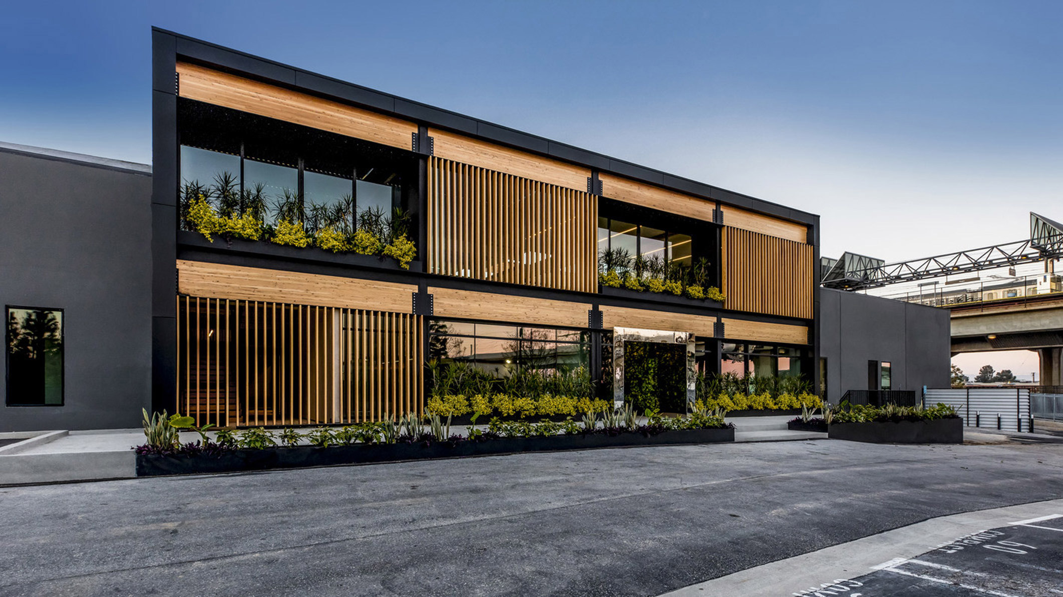 Modern commercial building exterior featuring sleek horizontal lines, wooden slat accents, and an integrated green space with planters on the upper level, captured during twilight hours.