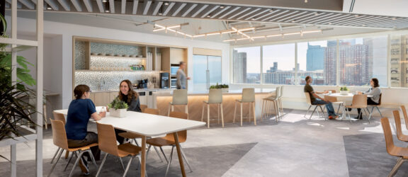Modern office break room with natural wood tables, light gray flooring, and suspended linear LED lighting. A kitchenette with mosaic backsplash and high stools overlooks a cityscape through large windows. Employees casually engage, fostering a collaborative atmosphere.