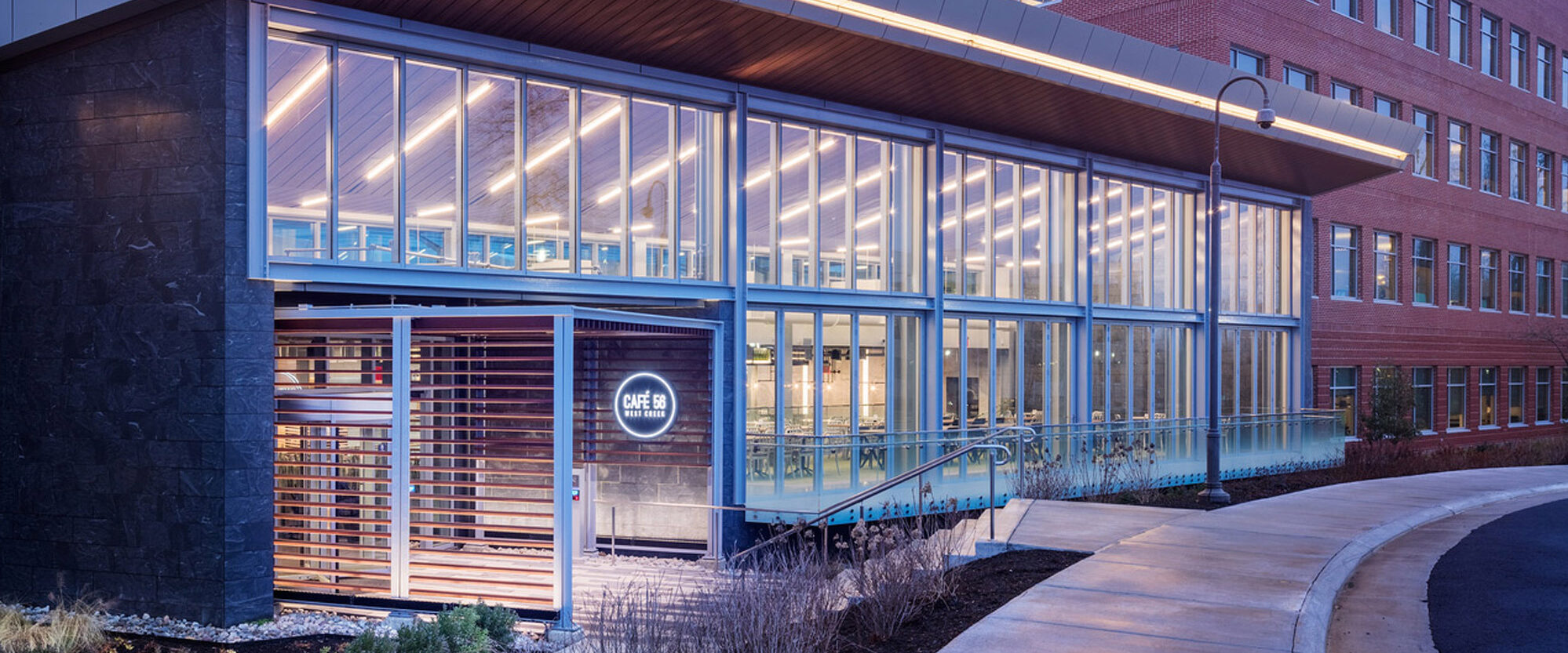 Modern educational building facade at twilight featuring a glass-walled walkway, illuminated interior spaces, sleek metal roof accents, and warm wooden cladding on a revolving entrance door area. The design integrates clear and opaque materials harmoniously, showcasing contemporary architectural style.
