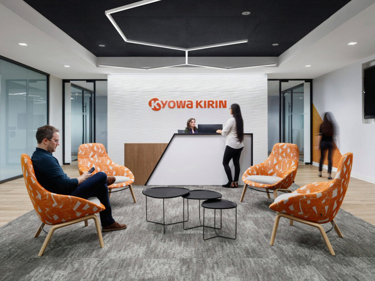 Modern corporate reception area featuring bold Kyowa Kirin brand wall, pops of orange in the upholstered armchairs, sleek black center tables, and dynamic lighting overhead, creating a welcoming ambiance for professionals and guests.