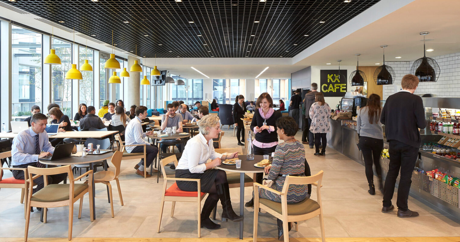Spacious café interior featuring a mix of communal and individual seating arrangements. Modern lighting with bright yellow pendants illuminates the space, while a sleek black grid ceiling adds a contemporary feel. Patrons engage at counters and tables in a bustling yet inviting atmosphere.
