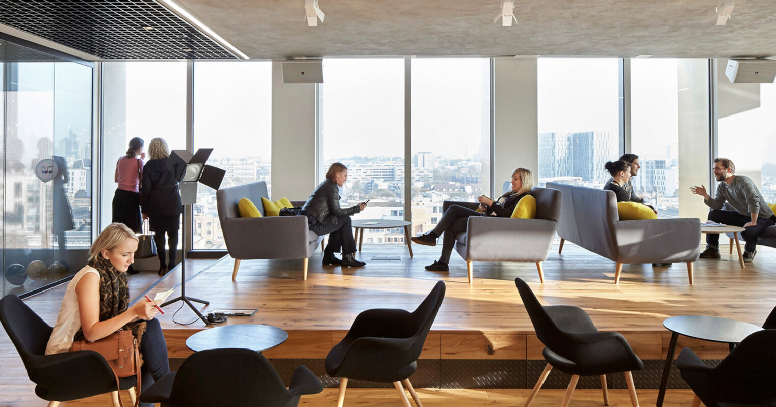 Contemporary office lounge with floor-to-ceiling windows offering a panoramic city view. The space features a mix of gray and yellow seating with sleek wood coffee tables, set on a natural wood floor and under an industrial-style black ceiling grid.