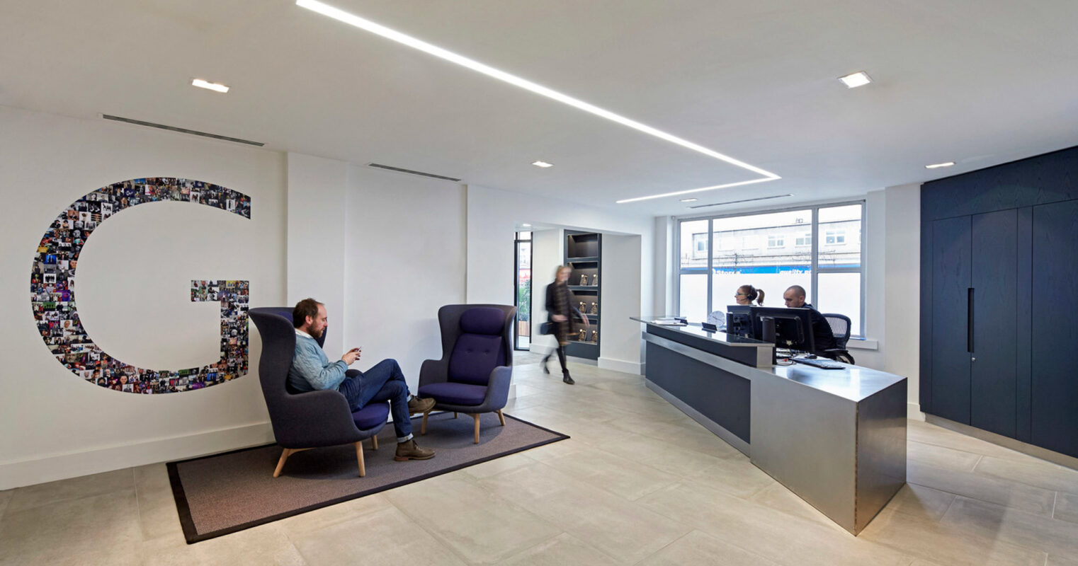 Modern office lobby with polished concrete floors, leading to a sleek reception desk. A prominent, mosaic-style company logo adorns the wall, complemented by recessed ceiling lighting and contemporary armchairs for waiting visitors.