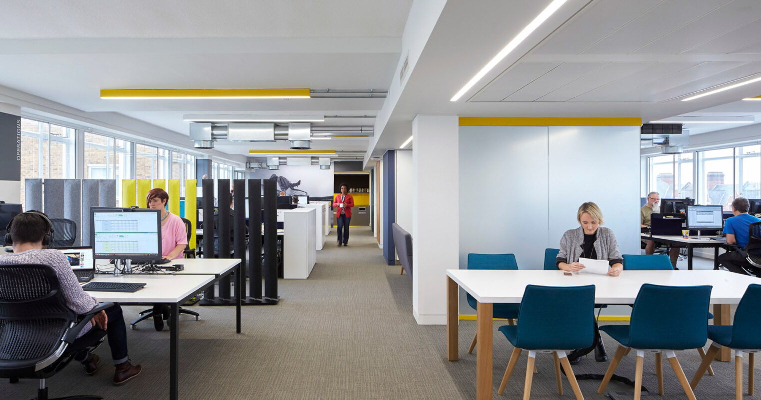 Modern office space featuring minimalistic workstations with white desks and ergonomic chairs. The open-plan area is punctuated by colorful acoustic panels, offering privacy and sound management. Daylight complements the LED strip lighting, casting a warm ambiance across the productive environment.