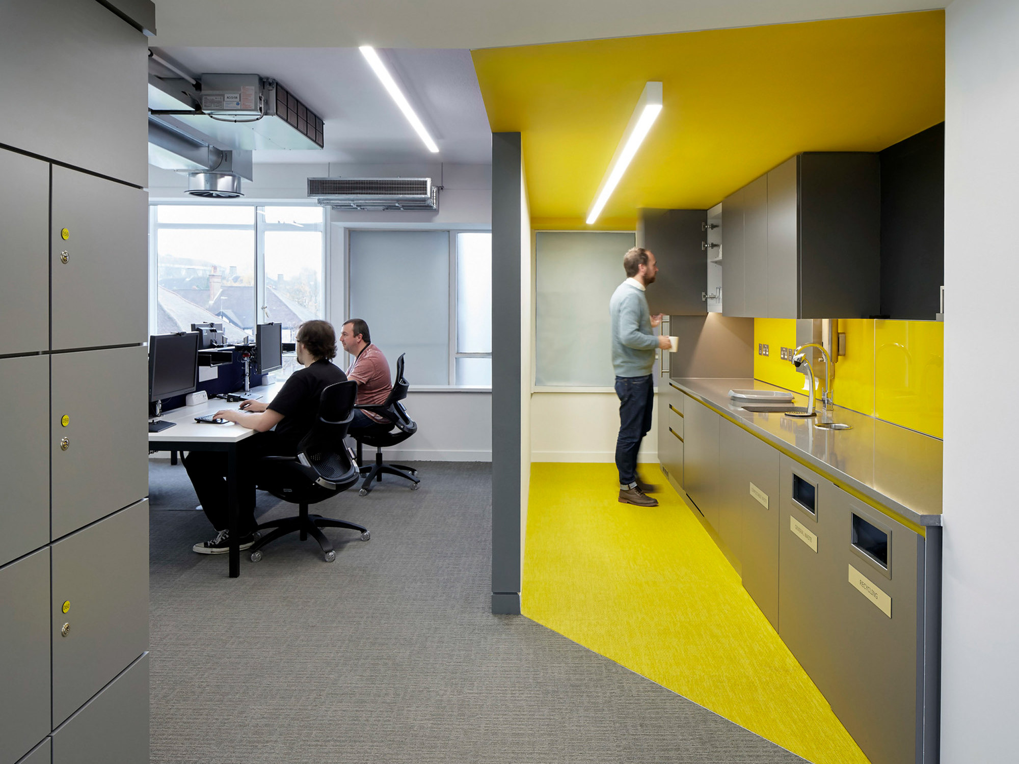 Modern office kitchenette with vibrant yellow backsplash and flooring, contrasting gray cabinetry, and stainless steel appliances. Two individuals work at adjacent desks, highlighting the space's functional and collaborative design.