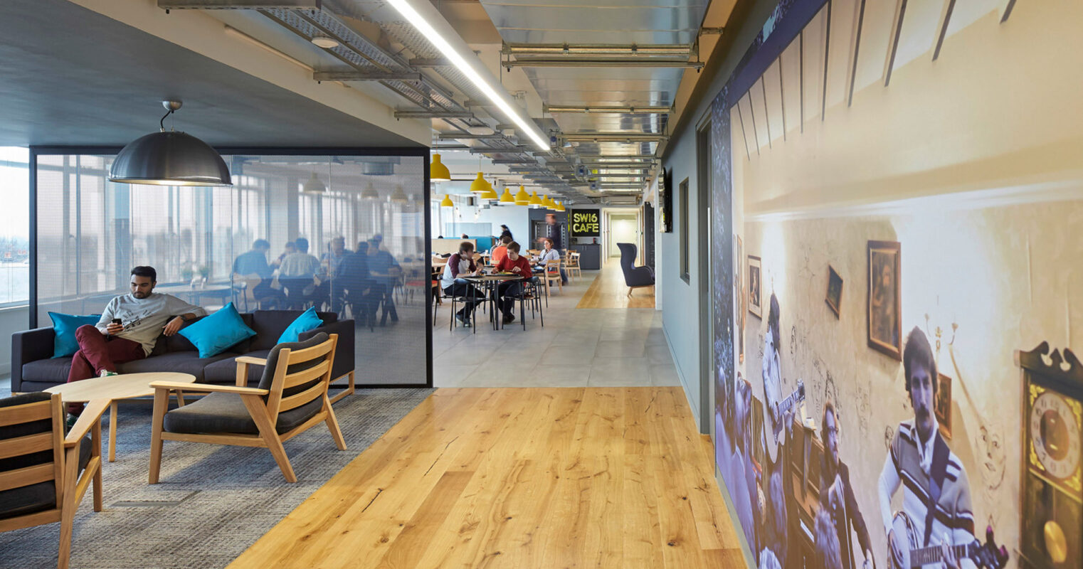 Modern office interior showcasing an open-plan layout with natural wood floors. Employees engage in collaborative work at colorful tables under yellow pendant lights. To the right, a casual seating area with a grey sofa provides a relaxed space beside a large historical mural.