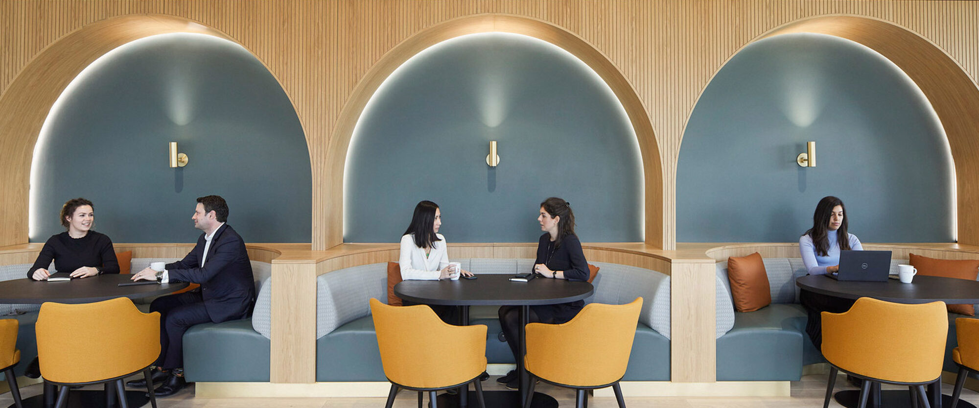 Contemporary office breakout area featuring teal arched alcoves with built-in seating, walnut wood accents, and mustard upholstered chairs. Individuals are casually engaged in work and conversation, affirming the space's collaborative design intent.