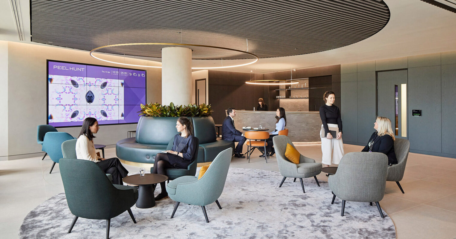 Modern office lounge with curved, modular seating in teal and charcoal. A circular ceiling feature complements the furniture's geometry, while a grey patterned rug anchors the space. Individuals engage in discussion, and a digital display animates the backdrop.
