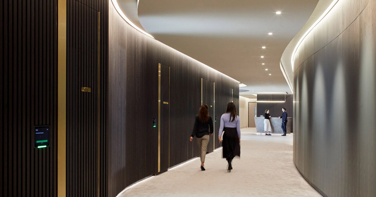 Curved hallway in a modern building with wood-paneled walls and recessed lighting, leading to a bright, open space where several individuals congregate.