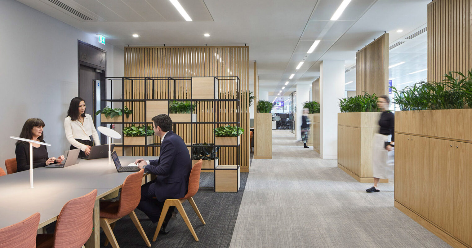 Modern office space featuring an open floor layout with sleek, white tables and coral chairs. Wooden slatted room dividers add texture, while employees engage in collaborative work under warm, ambient lighting.