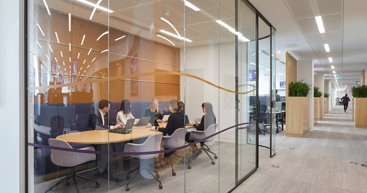 Modern office interior with open plan layout showcases clear glass partitions, wood veneer panels, and a streamlined oval conference table. Dynamic lighting accents above complement the natural light, while a collaborative meeting unfolds in the functional and stylish space.