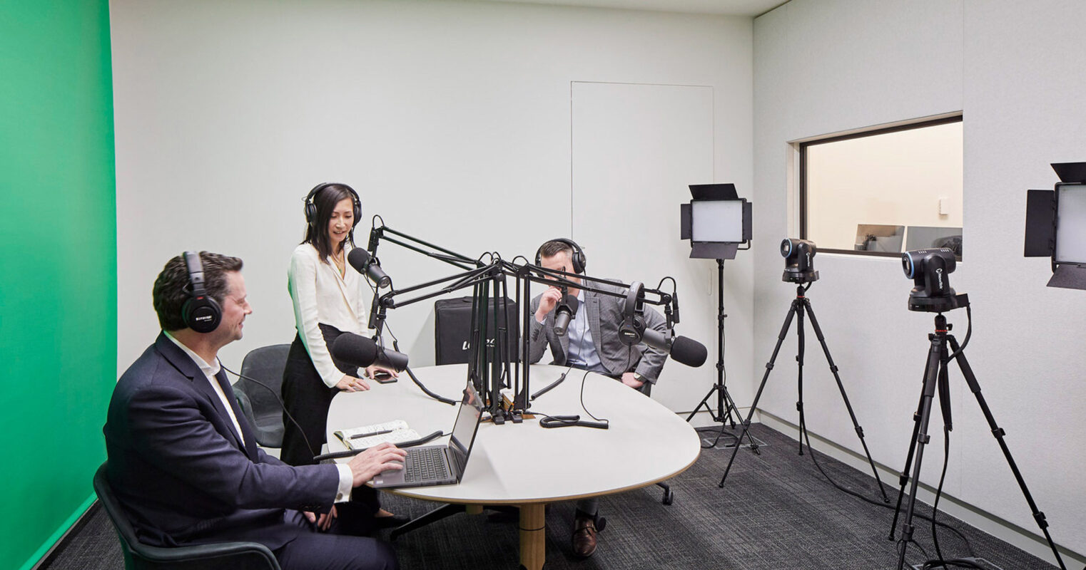 Minimalist podcast studio design with a neutral color palette, featuring soundproofed walls, modern broadcasting equipment, and ergonomic seating for three hosts. A glass partition separates the recording area, enhancing openness while maintaining acoustic privacy.