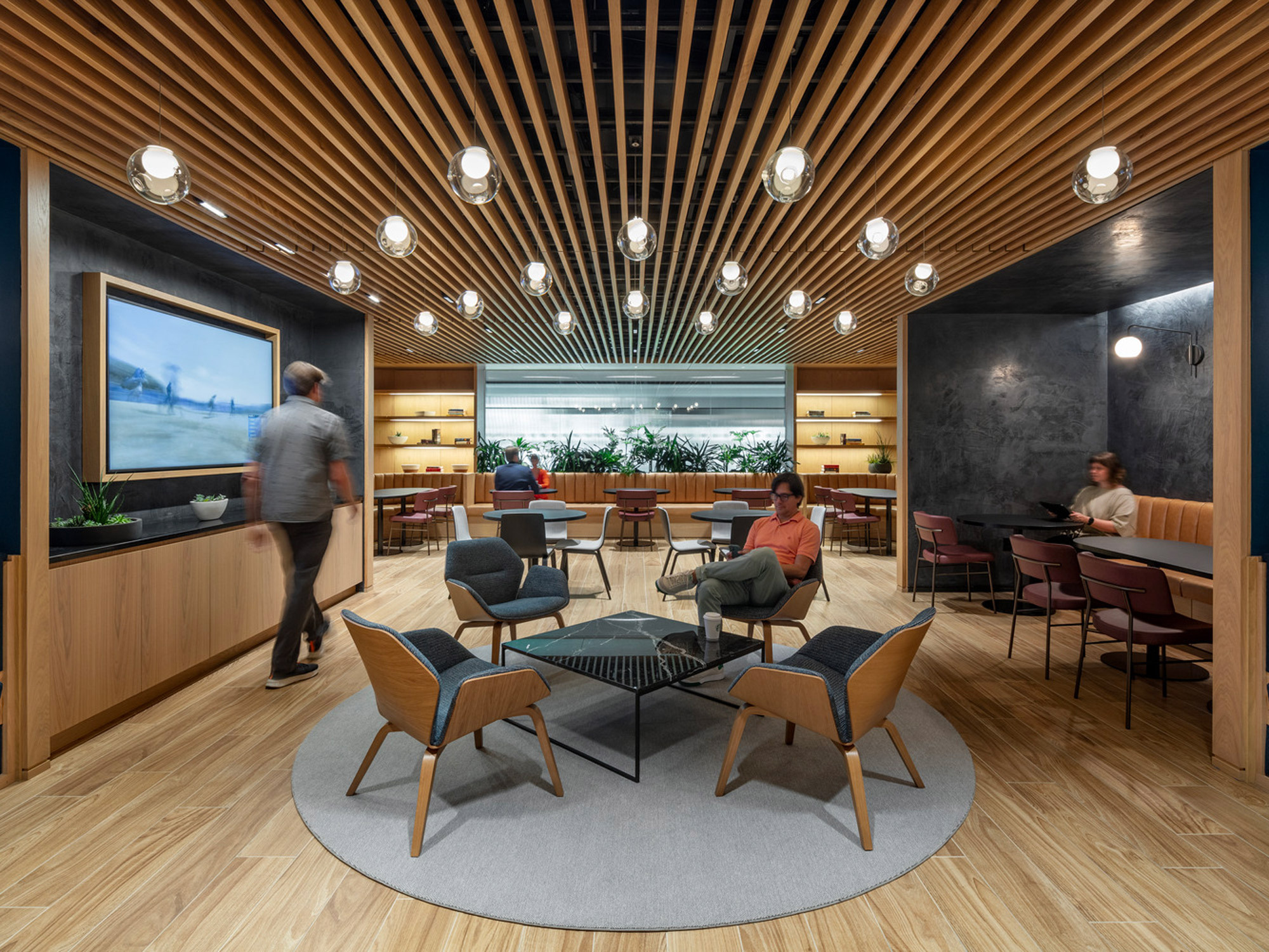 Modern office lounge with slatted wood ceiling and ambient globe lighting, complemented by mid-century-inspired furnishings, against a plant-filled window backdrop and textured dark wall accents.