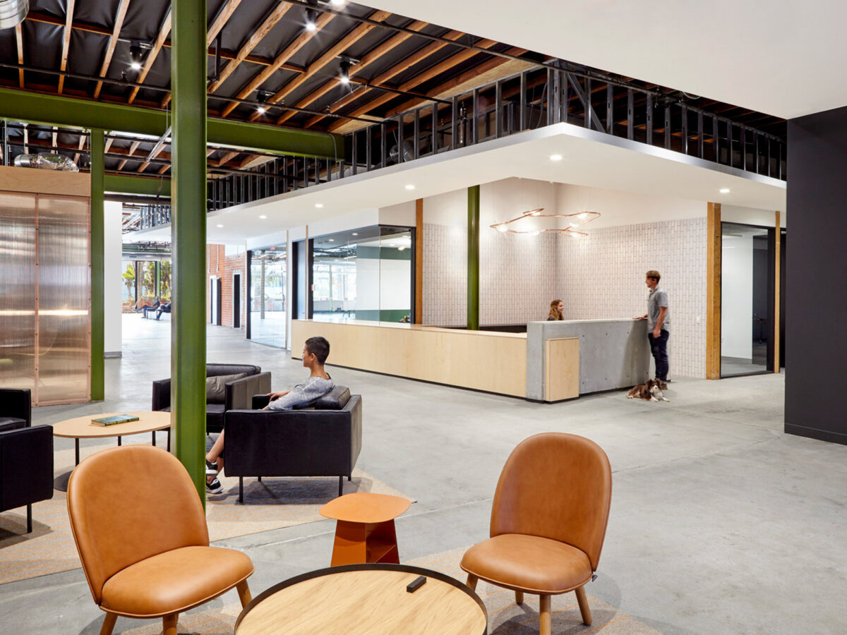 Modern office lounge with exposed ceiling beams and concrete pillars painted green. A minimalist reception desk is paired with sleek lighting fixtures, while tan leather chairs add a touch of warmth to the industrial ambiance.