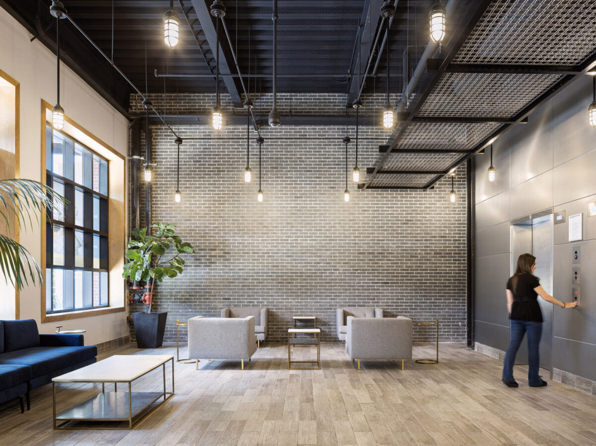 Modern industrial-inspired lobby with exposed brick walls, black ceiling with sleek track lighting, and polished concrete flooring. A minimalist gray sofa set complements the gold-framed glass coffee table, creating a sophisticated waiting area. Natural light streams in from large windows adding warmth.