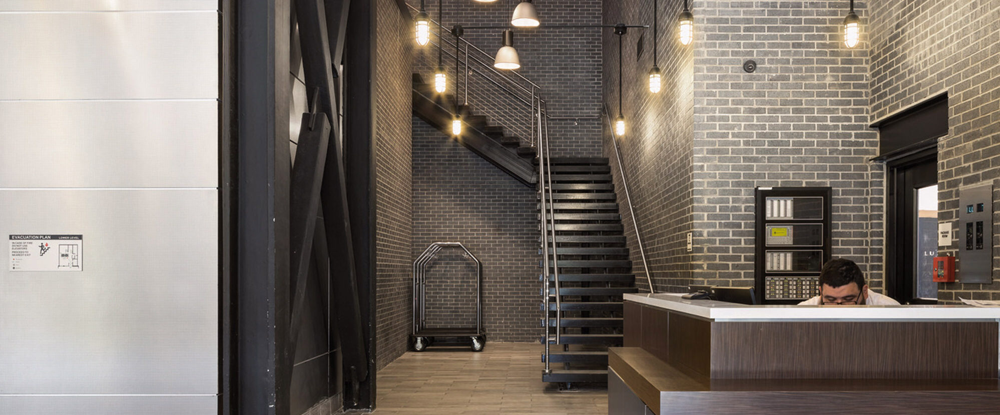 Modern industrial-style lobby featuring exposed black ceiling beams, brick walls, and pendant lighting. A sleek reception desk anchors the space while a metal staircase with cable railings leads upwards, complementing the dark wood flooring.