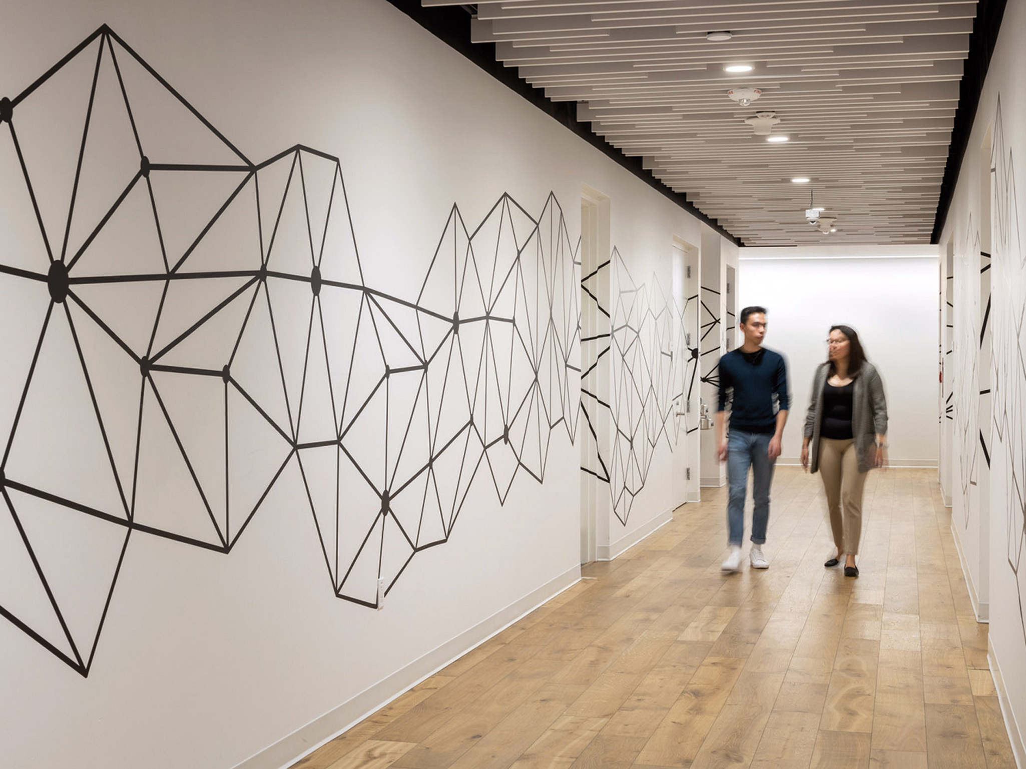 An expansive modern hallway with a geometric mural on the left wall, featuring abstract black line art on a white background. Linear lighting fixtures parallel the mural above, complementing the natural wooden flooring and minimalistic design as two individuals walk through.