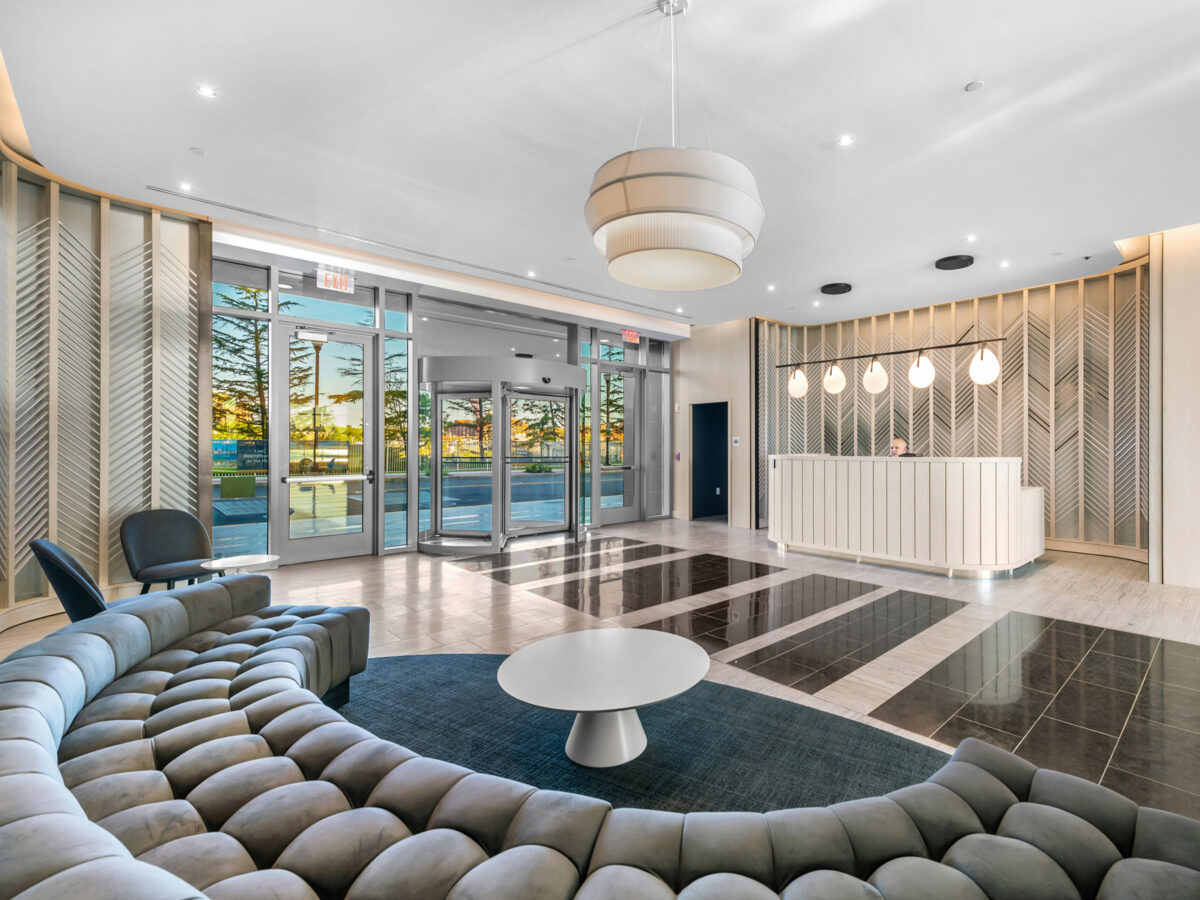 Modern hotel lobby featuring contrasting geometric floor patterns, tufted modular seating, a minimalist white reception desk, large hanging pendant lights, and floor-to-ceiling windows with warm natural light.