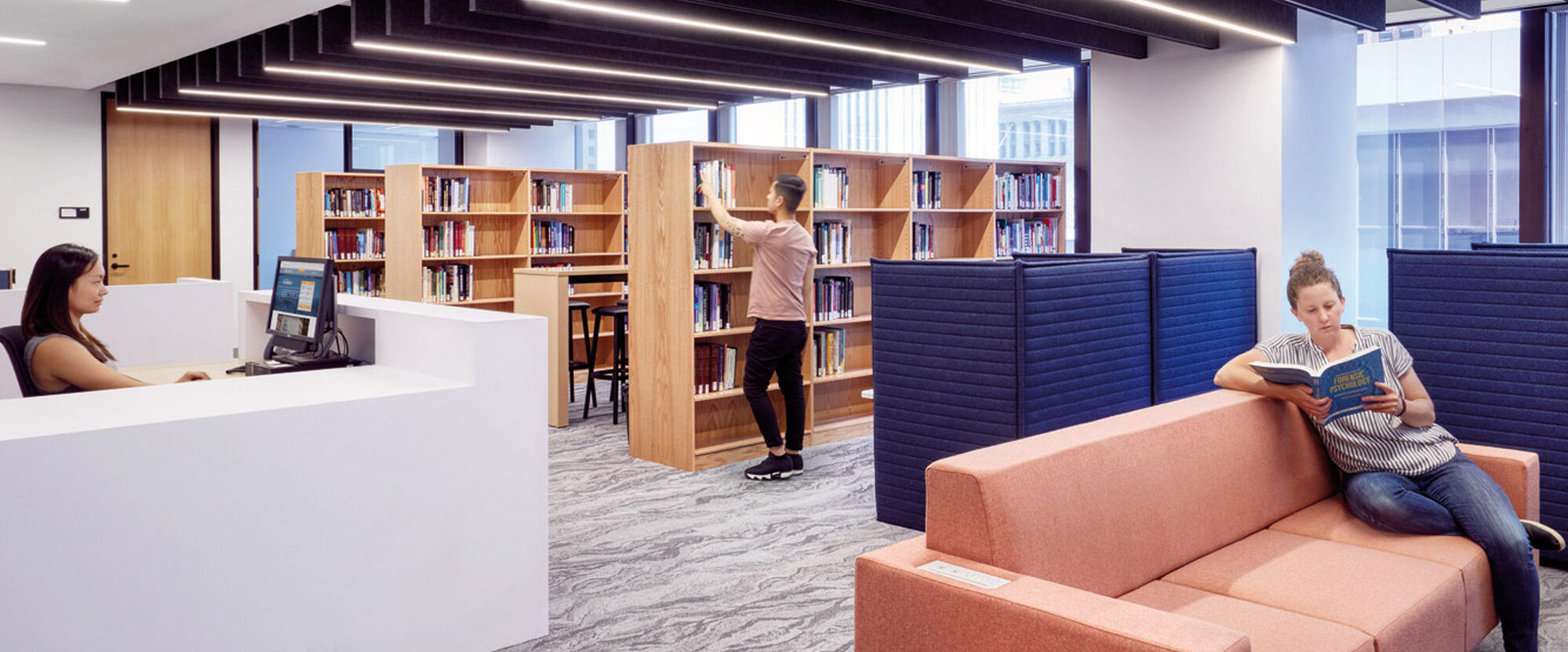 Contemporary library interior featuring streamlined white reception desk, peach-colored modular seating, and blue privacy panels. Dark ceiling with recessed lighting contrasts with the gray carpeted floors. The space is organized with sleek bookshelves, offering a quiet, modern reading environment.